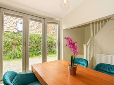 A dining area with a wooden table pink flowers in a pot teal chairs and glass doors showing a garden at Daisys Place in South Creake near Little Walsingham