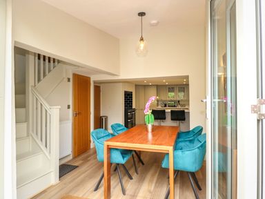 A dining area with a wooden table surrounded by teal chairs a staircase wooden doors and a kitchen with black stools at a counter at Daisys Place in South Creake near Little Walsingham