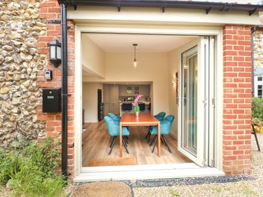 A dining area with a wooden table and blue chairs visible through open folding glass doors at Daisy's Place in South Creake near Little Walsingham
