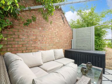 An outdoor seating area with a wicker sofa with cushions glass table with decorative items against a brick wall at Daisys Place in South Creake near Little Walsingham