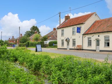 A street with a cream building with a red tiled roof and a bench outside with green plants and a small water channel in the foreground at Daisys Place in South Creake near Little Walsingham