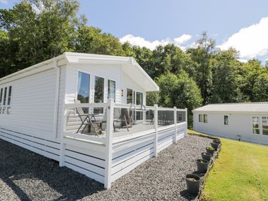 A white mobile home with chairs on a deck and a row of plant pots along a gravel path at Bethany's Retreat in Wemyss Bay