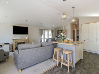 An open living area with grey sofas a kitchen island with two wooden stools and a television mounted above a fireplace at Bethany's Retreat in Wemyss Bay