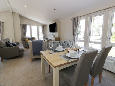 A combined dining and living area with a wooden dining table set with dishes and glasses near sofas and armchairs by windows at Bethany's Retreat in Wemyss Bay