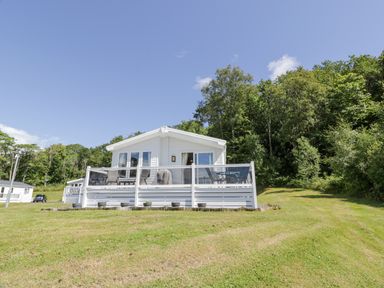 A white cottage with a deck and outdoor furniture on a lawn with trees in the background at Bethany's Retreat in Wemyss Bay