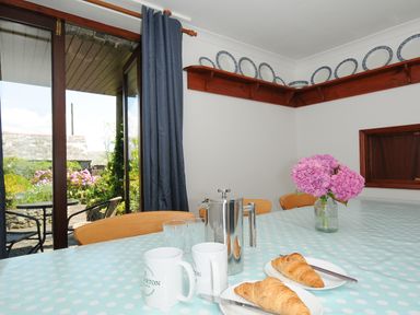 A dining room table set with croissants plates mugs coffee press and flowers near a glass door opening to a garden at West Lee in Bude