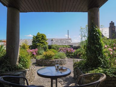 A patio area with a round glass table, two chairs, plants, flowers, and a stone building in the background at West Lee in Bude