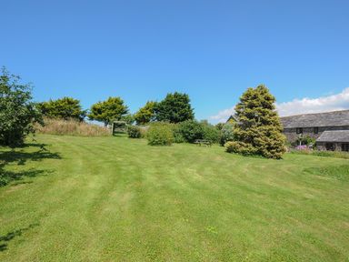 A grassy lawn area with trees bushes a wooden gate and a stone building with a slate roof at West Lee in Bude