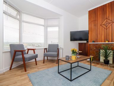 A sitting area with two grey chairs a wooden coffee table with mugs and flowers a wall-mounted TV and a fireplace at Small Hillman in Perranporth