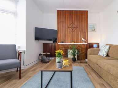 A living room with a wooden coffee table with mugs and a flower pot a TV mounted on the wall and a plant by a fireplace at Small Hillman in Perranporth