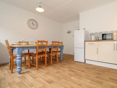 A dining area with a wooden table and chairs next to a white refrigerator and kitchen cabinets at Small Hillman in Perranporth