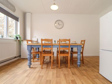 A dining area with a wooden table and six chairs a clock on the wall and a window with a plant at Small Hillman in Perranporth