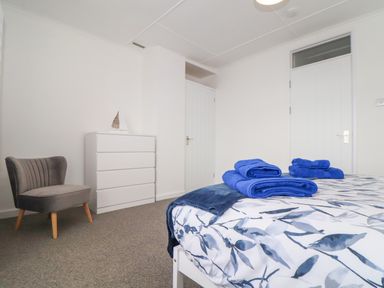 A bedroom with a bed covered in blue and white patterned bedding and folded blue towels beside a gray chair and white chest of drawers at Small Hillman in Perranporth