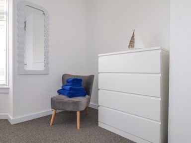 A grey chair with blue towels stacked on it next to a white dresser with a decorative sailboat and a wavy framed mirror on the wall at Small Hillman in Perranporth