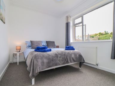 A bedroom with a double bed gray blanket and blue towels on the bed a bedside table with a lamp and a large window at Small Hillman in Perranporth