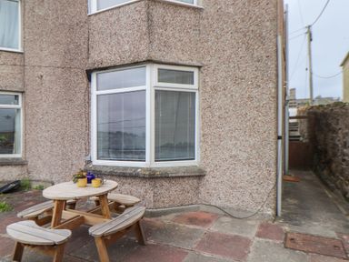 An outdoor patio area with a wooden picnic table and bench seating next to a house with pebble dash walls and bay windows at Small Hillman in Perranporth