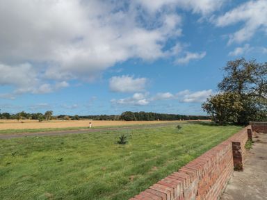 A grassy area with a brick wall and a rural field with trees under a partly cloudy sky at Curlew Cottage near Filey