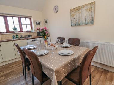 A dining area with a table covered with a polka dot tablecloth set with plates glasses flowers and a bottle near a kitchen at Curlew Cottage near Filey