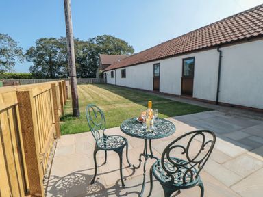 A patio with two metal chairs and a table with a bottle and glasses next to a lawn and a building with a tiled roof at Curlew Cottage near Filey