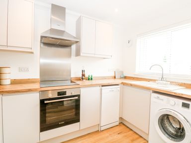 A kitchen with white cabinets wooden countertops stainless steel oven and extractor fan dishwasher washing machine and a window with blinds at Pine Cottage in Wrentham
