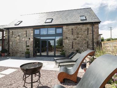 An outdoor patio area with modern lounge chairs a barbecue grill and a stone building with large glass doors at The Hay Barn in Huntley