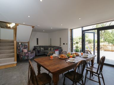A dining area with wooden table and chairs and a living area with sofa and armchair next to stairs and glass doors at The Hay Barn in Huntley