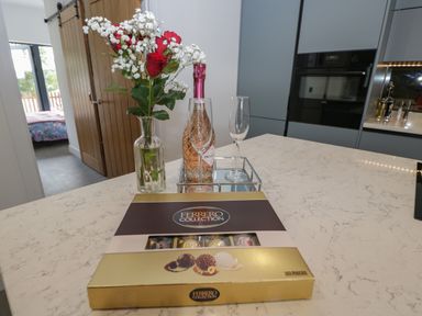 A kitchen countertop with a vase of red and white flowers a bottle of prosecco two glasses and a box of Ferrero Collection chocolates at The Hay Barn in Huntley