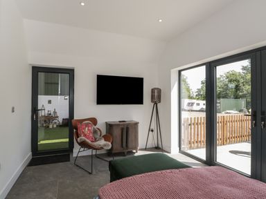 A living area with a wall-mounted TV brown chair with a pillow and throw a cabinet and a floor lamp next to glass doors at The Hay Barn in Huntley