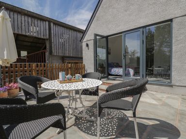 An outdoor patio with a white metal table, four black wicker chairs, a wooden fence, and open glass doors showing a bed inside at The Hay Barn in Huntley