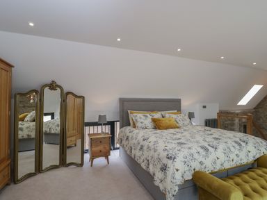 A bedroom with a large bed floral bedding a wooden bedside table with a lamp a three-panel mirror a wooden wardrobe and a green upholstered bench at The Hay Barn in Huntley