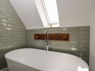 A bathroom with a white bathtub green tiled walls a skylight and a recessed wooden shelf at The Hay Barn in Huntley