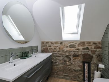 A bathroom with a double sink vanity a round mirror a stone wall and a skylight window at The Hay Barn in Huntley