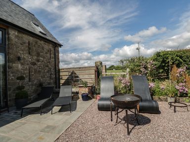 An outdoor patio area with four lounge chairs a fire pit stone wall and plants at The Hay Barn in Huntley