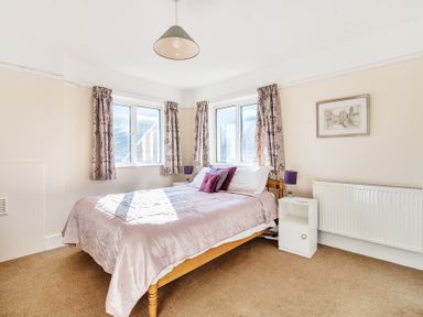 A bedroom with a double bed pink bedding purple cushions two windows with floral curtains bedside tables with purple lamps a framed picture on the wall and a radiator at Bayview in Overcombe near Preston