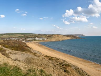 A sandy beach with a few people walking near a grassy hillside and buildings along the coast at Bayview in Overcombe near Preston