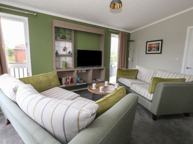 A living room with two sofas, a wooden coffee table with cups, a wall-mounted shelving unit with a TV and decorative items at Hazelwood 2 Pinewood near Lyme Regis