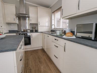 A kitchen with white cabinets gray countertops a stove a microwave bread on a cutting board and a window with blinds at Hazelwood 2 in Pinewood near Lyme Regis