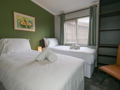 A bedroom with two single beds with rolled towels on each bed a window with curtains and blinds a green accent wall with a painting a brown chair and a shelving unit at Hazelwood 2 in Pinewood near Lyme Regis