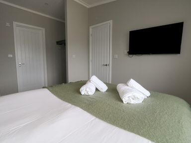 A bedroom with a bed covered in white and green linens with rolled white towels on top a wall-mounted TV and two white paneled doors at Hazelwood 2 Pinewood near Lyme Regis
