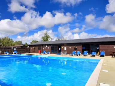 An outdoor swimming pool with blue lounge chairs and a brown building in the background at Hazelwood 2 Pinewood near Lyme Regis