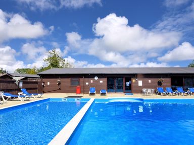 An outdoor swimming pool with blue lounge chairs and a brown building in the background at Hazelwood 2 Pinewood near Lyme Regis