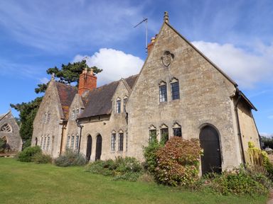 Exterior view of a stone building with gothic windows and chimneys surrounded by greenery at The Alms House East in Forthampton near Tewkesbury