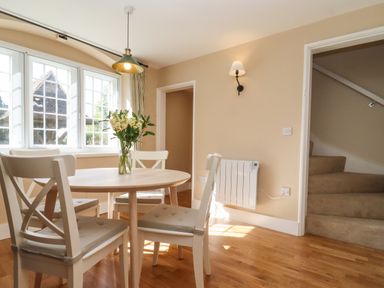 A dining area with a round wooden table and four chairs near a large window and stairs at The Alms House East in Forthampton near Tewkesbury