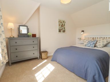 A bedroom with a bed with blue bedding and patterned cushions a grey dresser with a lamp mirror and decor a window and a framed bird picture at The Alms House East in Forthampton near Tewkesbury