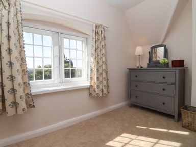 A room with a window with floral curtains and a gray dresser with a lamp mirror and basket at The Alms House East in Forthampton near Tewkesbury