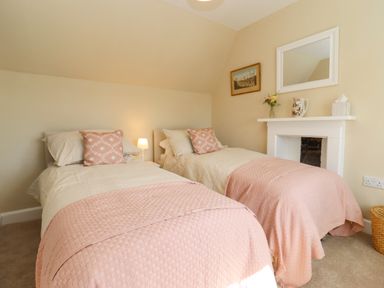 A bedroom with two single beds with pink blankets and pillows a small lamp on a nightstand and a white mantle with a mirror a jug flowers and tissues at The Alms House East in Forthampton near Tewkesbury