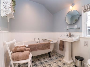 A bathroom with a clawfoot bathtub a pedestal sink round mirror towels on a chair and patterned floor tiles at The Alms House East Forthampton near Tewkesbury