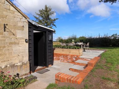 An outdoor patio area with a wooden table and chairs next to a stone wall and black door at The Alms House East in Forthampton near Tewkesbury