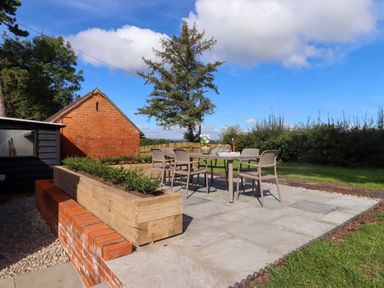 An outdoor patio with a table and six chairs surrounded by a wooden planter and brick wall at The Alms House East in Forthampton near Tewkesbury
