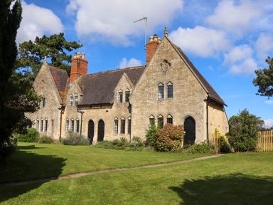An old stone house with multiple chimneys and arched windows surrounded by grass and bushes at The Alms House East in Forthampton near Tewkesbury